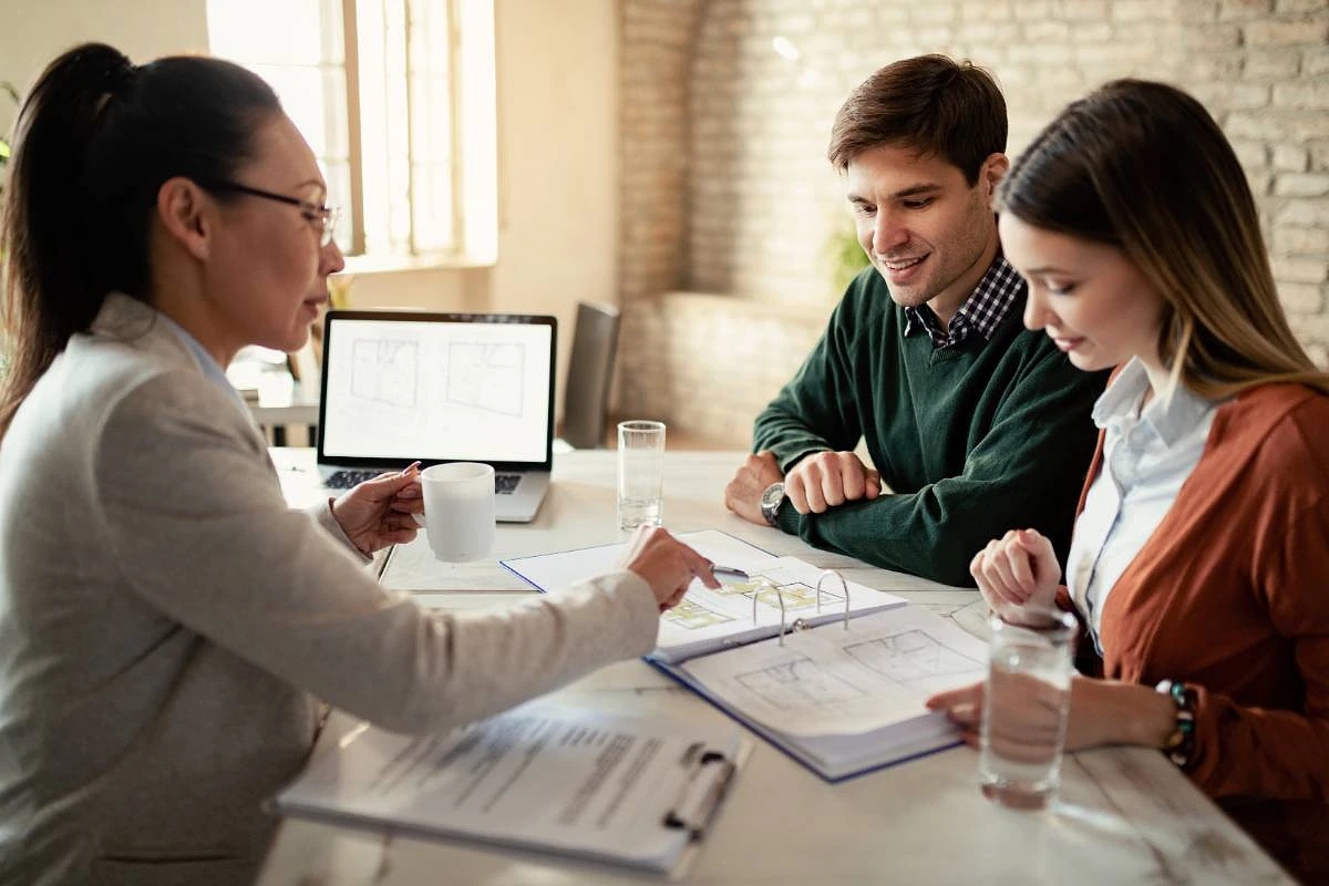 Three people are having a meeting at a table with documents and a laptop displaying floor plans.