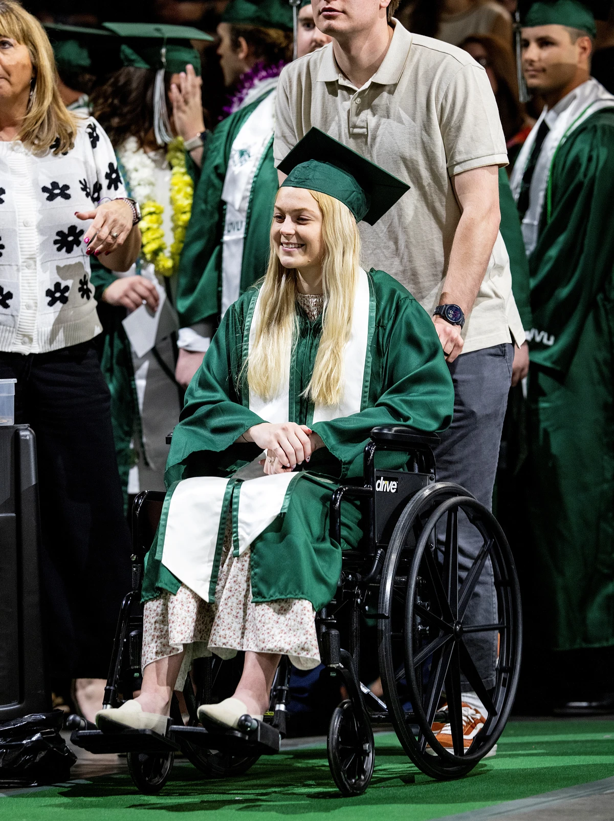 A woman in a wheelchair, wearing a green graduation gown and cap, smiles while surrounded by others at a graduation ceremony.