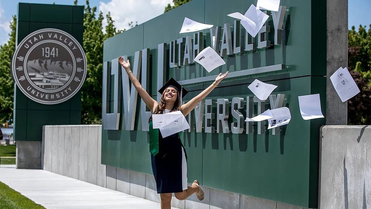 A graduate joyfully throws papers into the air in front of the Utah Valley University sign.